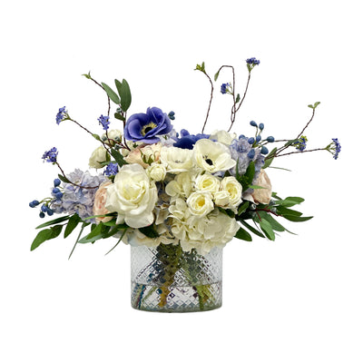 Hand-crafted realistic floral arrangement with lifelike delphinium, hydrangea, and delicate accent blooms, presented in an elegant cut glass vase, placed against a white background.